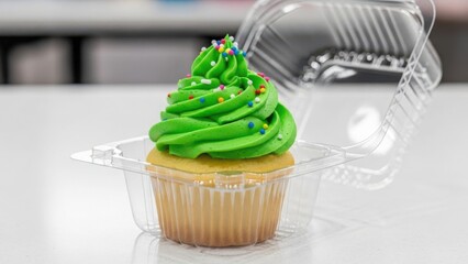 A single cupcake with bright green frosting and colorful sprinkles in a plastic to-go container. Sweet dessert for a birthday party or celebration