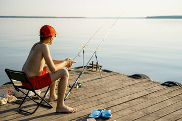 Young boy relaxes on a dock by a tranquil lake, enjoying a peaceful fishing session. Wearing a cap and shorts, he holds fishing rods, immersed in the calming outdoor atmosphere.
