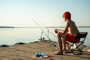Young boy in red shorts and cap fishing on a wooden dock during a summer day. He is sitting on a foldable chair, holding fishing equipment, surrounded by calm water and blue sky.