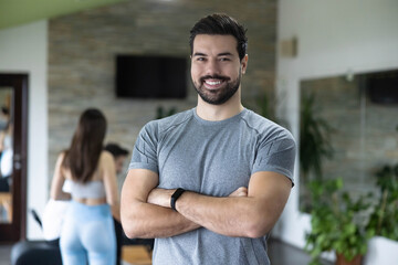 A confident young man stands with his arms crossed in a pilates studio, showcasing his fitness while exuding a friendly and approachable vibe, perfect for fitness promotions.