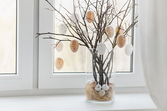 Creative Easter home decor with beige pastel eggs and tree branches in glass vase on windowsill. Bright spring atmosphere, natural light. Home interior.