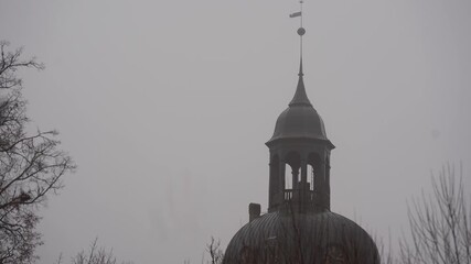Towering dome structure with a weather vane is partially obscured by fog, showcasing architectural details and surrounding trees in a misty atmosphere