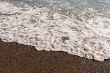Water meets sand as waves roll onto the shore in a warm coastal location. Light from the sun reflects off the water in the early afternoon