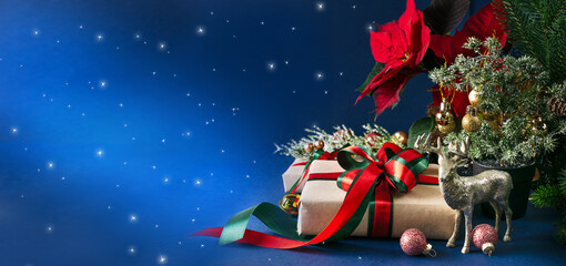 Christmas banner with gift box with bow, red poinsettia, decorative fir branch, deer and balls for tree decoraton on blue wide background. New Year winter holiday composition with decor and presents.