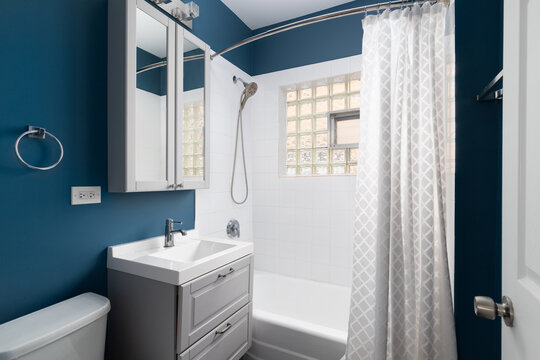 A blue bathroom featuring a grey vanity, white sink, and white square tiled walls with a glass block window.