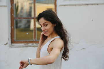 Happy beauty face woman looking on the time on watch on hand with long brown hair in casual clothes outdoor summer houses window background. Closeup fun relaxing person street portrait