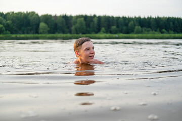 Fototapeta premium A young boy enjoying a swim in a calm lake, surrounded by lush green trees. His expression reflects joy and relaxation as he engages with nature during a peaceful, sunny day.