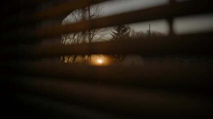 Warm sunset light shines through partially closed window blinds, creating soft, moody atmosphere inside the room. Person opening the blinds - Powered by Adobe