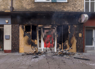 Burned storefront facade after a fire, showing severe fire damage, broken windows, charred walls, and debris on the sidewalk.