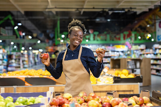 Happy african american grocery worker wearing an apron, smiling while holding fresh red and green apples by a vibrant produce display in a supermarket, conveying friendly service and healthy options - Powered by Adobe