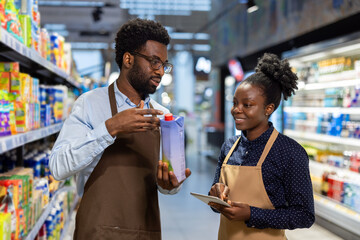 Young man and woman, both employees wearing aprons, coordinating and discussing product inventory while using a digital tablet in a grocery store aisle