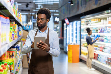 Supermarket employee in an apron and glasses standing in an aisle, carefully checking product inventory on a digital tablet while another worker stocks shelves in the background