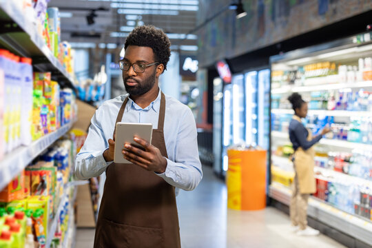 Supermarket employee in an apron and glasses standing in an aisle, carefully checking product inventory on a digital tablet while another worker stocks shelves in the background