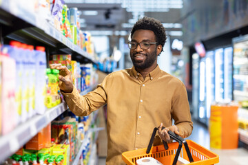 Happy african american man choosing products from a shelf in a supermarket aisle, holding an orange...