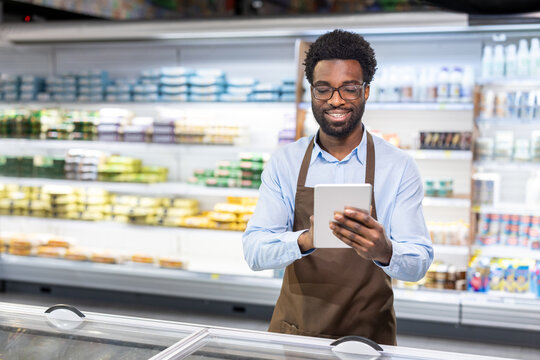 Young african american male supermarket employee wearing an apron and eyeglasses, smiling and checking inventory on a digital tablet in a grocery store aisle - Powered by Adobe