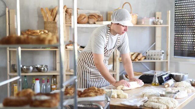 Young guy working at bakery rolling out dough into sausages, young male baker kneading dough and shaping baguettes on countertop in small kitchen 