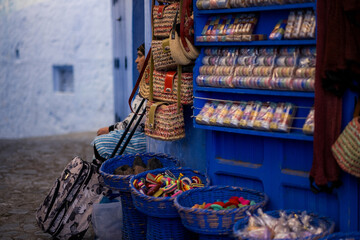 Colorful woven baskets and handmade goods line a blue-painted shop wall in Chefchaouen, while a local woman rests nearby, capturing the calm rhythm of everyday life in the blue medina.