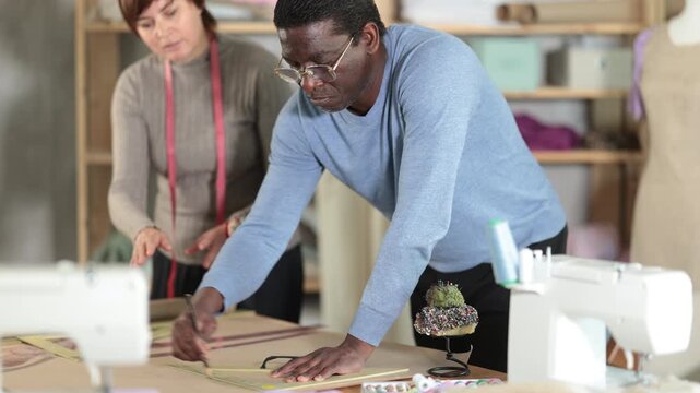 Woman tailor draw dress pattern on paper and participate in creating pattern with African man companion. Craftsman perform paper prototype of product parts, drawing paper pattern for dressmaking.