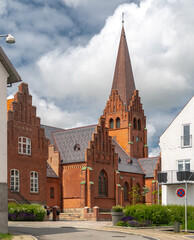 Historic red-brick church in Nykøbing Mors, North Jutland Region, Denmark—steep spire, stepped gables, and Gothic Revival details framed by modern buildings under a bright summer sky © uslatar