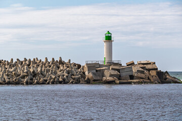 Green lighthouse standing on a concrete breakwater made of large wave-breaking blocks, with calm sea and sky in the backgroun