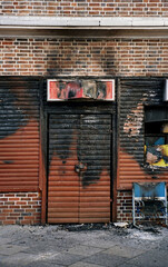 Burned and vandalized kiosk facade with charred wooden shutters and fire-damaged brick wall in an urban environment.