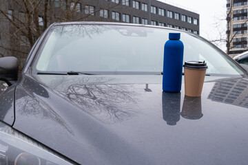 Thermos bottle and paper coffee cup standing on car hood