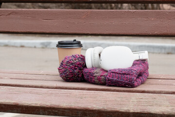 Takeaway coffee cup, cozy knit hat, and wireless headphones lying on a wooden bench