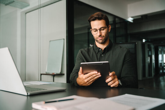 Focused Latin entrepreneur businessman thinking holding digital pc tablet sitting in office. European young man working on trade online using touchpad fintech computer for business work. Copy space