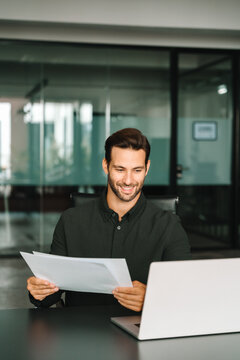 Smiling latin business man salesman working on laptop computer reading financial contract document in office workplace. Young manager businessman specialist doing paperwork analysing on pc. Vertical