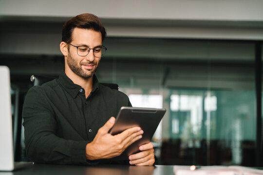 Smiling European 30s trade manager looking at digital tablet screen sitting at desk in office. Executive professional man working online using computer for financial business banking work. Copy space