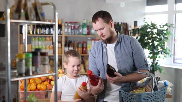 Father with boy hold packages plastic bottle and choose consider fruit juice in store. Market visitors choose make choice and view bottle juice. Client hesitates before buying, chooses drink