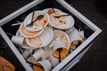 Overflowing trash bin filled with used disposable plates, cups, and utensils, showcasing the aftermath of a busy outdoor event with food remnants and waste materials