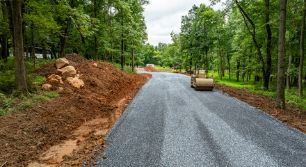 Workers are constructing a gravel road in a wooded area. Heavy machinery is on site, moving dirt and laying down gravel. The sky is cloudy and the ground is uneven with rocks and dirt piles.