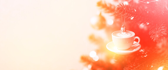 Close-up shot of white coffee cup ornament with latte art hanging on Christmas tree branch with warm bokeh.