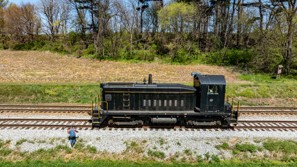 A black locomotive traverses rail tracks while a person stands beside it on the ground. The...