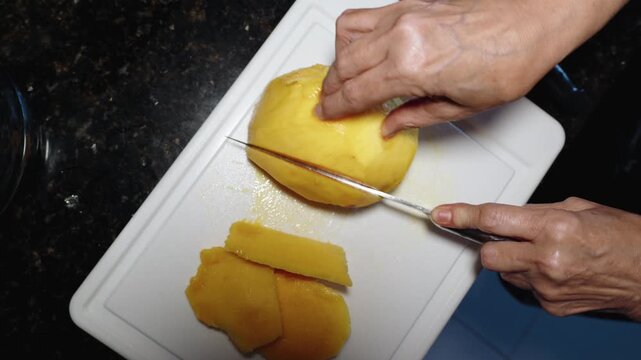 Top-down view of senior woman rheumatoid arthritis hands using a knife to slice a mango on a white cutting board
