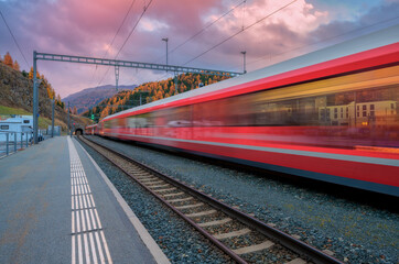 Blurred red passenger train passing mountain railway station in Swiss Alps at dusk. Moving high-speed train. St. Moritz, Switzerland. Bernina Express.  Railway platform and cloudy sky. Rail transport