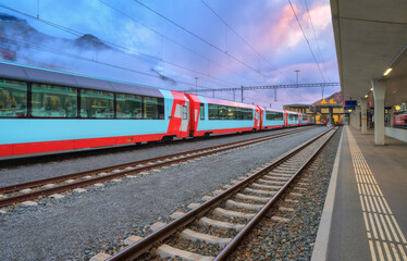 Red and blue modern passenger train standing at railway station in Swiss Alps at dusk. Mountains in fog, tracks and railway platform under purple cloudy sky. St. Moritz, Switzerland. High-speed train