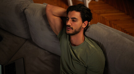 Obraz premium Young man relaxing comfortably on a gray sofa in his living room, casually watching television or a screen, unwinding in a comfortable atmosphere, enjoying leisure time