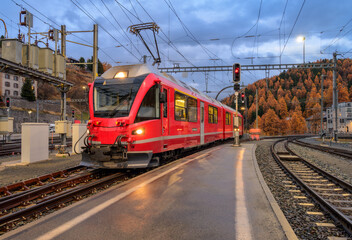 Red modern train on mountain railway station in Swiss Alps in autumn at dusk. Speed train with lights, railroad, rain, wet railway platform, orange trees in fall at dusk. Switzerland. Bernina Express
