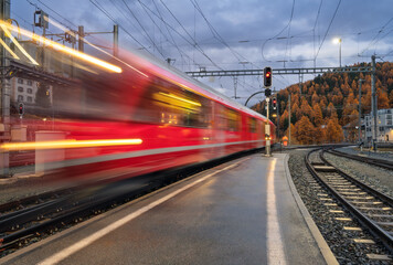 Blurred red passenger train passing mountain railway station during rain in Swiss Alps at night. Moving speed train at dusk. St. Moritz, Switzerland. Bernina Express.  Railway platform and lights.