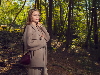 A glamour woman is standing in a forest, wearing a brown coat and holding a purse. The image has a natural and peaceful feel to it, with the woman posing in the woods surrounded by trees and greenery