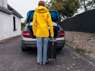 A woman in a yellow jacket is standing next to a car trunk with a suitcase. The scene is casual and relaxed, with the woman likely preparing for a trip. Travel and tourism concept.