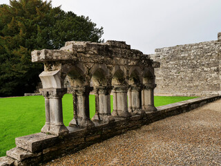 A stone wall with a row of pillars and a green grassy area in the background. The wall is old and the pillars are made of stone. The grass is lush and green, giving the scene a peaceful