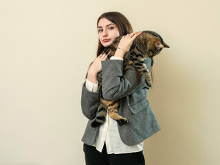 Portrait of a beautiful young business woman holding big cute tabby cat in her hands. Owner with he beloved pet. Light color background. Model with long dark hair in grey suit.