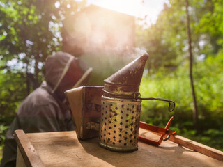 Old metal bee smoker on a wooden table, beekeepers out of focus in the background. Scene on a farm during beehive inspection and honey pick up.
