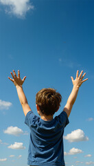 A boy stands in an open area with his arms raised high. He looks toward the bright sky with clouds. The sun is shining, and it is a clear day