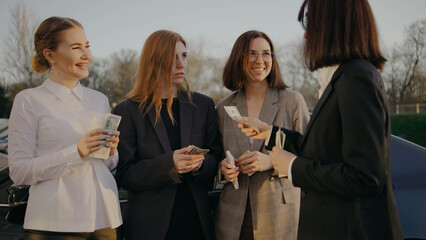 Group of women exchanging money in a parking lot during the late afternoon under clear skies with trees in the background
