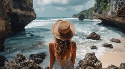 A woman stands on a beach looking out at the ocean. The scene includes rocky cliffs waves rolling in and a cloudy sky. It is daytime in Bali.