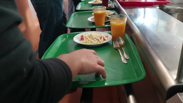 Customers slide trays along metal rails to select lunch dishes in a busy public dining room, showing a self-service cafeteria line and diverse meal options in high-quality 4k resolution.